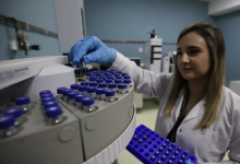 Bir sağlık çalışanı laboratuvarda beyaz önlük ve mavi eldivenlerle küçük numune tüplerini dikkatle yerleştiriyor. Önünde dairesel bir örnek tepsisi ve analiz cihazı bulunuyor; ortam steril ve profesyonel bir çalışma alanı hissi veriyor.