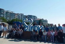 Görselde, “Mutlu Kent Sinop” yazılı büyük dekoratif yapının önünde bir grup insan dış mekânda toplu hâlde poz veriyor. Arkada apartmanlar ve ağaçlar görülüyor. Hava güneşli; ortam samimi, şehir meydanında çekilmiş bir hatıra fotoğrafı izlenimi veriyor.