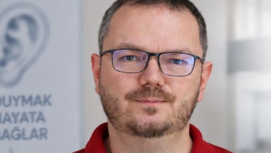 Görselin üst bölümünde, kırmızı polo yaka tişört giymiş, dikdörtgen koyu çerçeveli gözlük takan ve kirli sakallı bir erkek uzman hekimin doğrudan kameraya baktığı portresi yer alıyor. Arka planda aydınlık bir klinik veya ofis ortamı ile sol tarafta kulak çizimi ve "DUYMAK HAYATA BAĞLAR" sloganının bir kısmı okunan bilgilendirici dikey bir afiş görülüyor.
