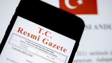 Görselde, yakın plan çekilmiş bir akıllı telefon ekranında T.C. Resmî Gazete ibaresi ve yayım detayları yer almaktadır; arka planda ise Türk bayrağı silüeti ve yazılı kağıtlar bulunmaktadır.