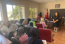 Görselde, aydınlık bir odada koltuklarda oturan katılımcı kadınlara sunum veya eğitim veren bir eğitmen yer almaktadır. Odanın köşesinde Türk bayrağı ve duvarda çerçeveli bir tablo bulunmaktadır.