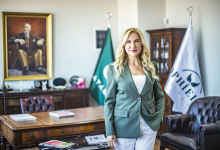 Dr. Şeyda Atabay, modern ve kurumsal bir ofis ortamında ayakta duran, profesyonel bir duruşa sahip bir yöneticidir. Açık renk saçları, beyaz üst ve pantolonla uyumlu yeşil bir blazer ile sade ama güçlü bir stil yansıtır. Arkasında kurum kimliğini gösteren bayraklar ve büyük bir portre bulunur. Çalışma alanı; ahşap masa, dergi ve kitaplar, deri koltuklar gibi detaylarla resmî, akademik ve yönetimsel bir atmosfer sunar. Genel görünümü; liderlik, temsil gücü ve kurumsal otorite hissi verir.