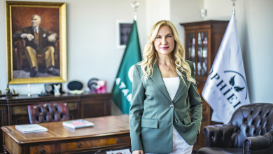 Dr. Şeyda Atabay, modern ve kurumsal bir ofis ortamında ayakta duran, profesyonel bir duruşa sahip bir yöneticidir. Açık renk saçları, beyaz üst ve pantolonla uyumlu yeşil bir blazer ile sade ama güçlü bir stil yansıtır. Arkasında kurum kimliğini gösteren bayraklar ve büyük bir portre bulunur. Çalışma alanı; ahşap masa, dergi ve kitaplar, deri koltuklar gibi detaylarla resmî, akademik ve yönetimsel bir atmosfer sunar. Genel görünümü; liderlik, temsil gücü ve kurumsal otorite hissi verir.