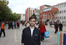 Görselin üst bölümünde, bir okulun bahçesinde, arkasında okul binası ve öğrenciler bulunan görme engelli genç bir öğretmen yer almaktadır. Siyah fermuarlı bir spor ceket giyen ve gözlük takan öğretmen, doğrudan kameraya bakarak hafifçe gülümsemektedir. Arka plandaki okul binasının üzerinde "Fatih Sultan Mehmet Ortaokulu" tabelası okunurken; bahçede okul formalı öğrenciler dolaşmakta, öğretmenler ve hareketli bir okul ortamı göze çarpmaktadır.