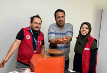Instagram İçin Alternatif Metin: Görselin üst bölümünde, çömlek atölyesinde elektrikli çömlek çarkı makinesinin arkasında yan yana durarak poz veren üç kişi yer alıyor. Ortada çizgili polo yaka tişört giymiş engelli birey, iki avucunun içinde kilden yaptığı minik çömlek kabını gururla ileriye uzatarak sergiliyor. Solunda kırmızı Gençlik ve Spor Bakanlığı yeleği giymiş, elleri kile bulanmış eğitmen genç adam; sağında ise koyu yeşil başörtülü ve aynı kırmızı yelekten giymiş gülümseyen genç bir kadın görevli duruyor. Önlerinde turuncu gövdeli döner çömlek çarkı ve açık renkli sade duvar arka planı görülüyor.