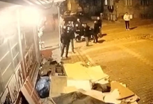 Görselde, gece vakti sokakta güvenlik kamerası açısıyla yukarıdan çekilmiş bir görüntü yer almaktadır. Bir binanın önünde veya dükkan kenarında toplanmış bir grup insan ve etrafta karton parçaları, çöpler görülmektedir.