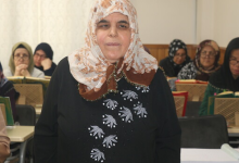 Görselde, kapalı bir alanda ön planda yer alan, desenli başörtülü ve koyu renk kıyafetli bir kadın ile arka planda masalarda oturarak ders çalışan veya kitap okuyan diğer katılımcılar yer almaktadır.