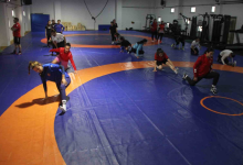 Görselin üst bölümünde, kapalı spor salonundaki mavi ve turuncu zeminli güreş minderi üzerinde antrenman yapan İşitme Engelli Kadın Güreş Milli Takımı sporcuları ve antrenörleri yer alıyor. Ön planda mavi ve kırmızı antrenman formaları giymiş kadın güreşçiler minderde teknik hareket ve taktik çalışması yaparken; orta alanda beyaz tişörtlü bir sporcu diğer takım arkadaşlarıyla ilgileniyor. Sol tarafta ise kırmızı tişörtlü ve siyah şortlu erkek antrenör kollarını kavuşturmuş vaziyette sporcuların çalışmasını dikkatle takip ediyor. Arka fonda mavi koruma minderleri, ahşap antrenman kasaları ve spor salonunun geniş iç mekânı görülüyor.