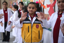Görselin üst bölümünde, beyaz doktor önlükleri giymiş işitme engelli öğrenciler yer alıyor. Merkezde duran saçları toplu kız çocuğu, beyaz önlüğünün üzerine sarı ve gri renkli acil sağlık/ambulans görevlisi yeleği giymiş olarak iki elinin avuç içini ileriye doğru açarak işaret diliyle poz veriyor. Sağ ve sol tarafta beyaz önlük giymiş diğer kız ve erkek öğrenciler de ellerini kaldırarak işaret dilinde iletişime katılıyor. Arka planda renkli çizimlerin ve el ele tutuşan figürlerin bulunduğu bir duvar panosu görülüyor.