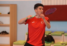Görselin üst bölümünde, kapalı bir spor salonunda kırmızı-bordolu millî takım forması giymiş genç bir erkek badminton sporcusu elinde raketle vuruş pozisyonu almış halde yer almaktadır. Odaklanmış bir ifadeyle ileriye bakan sporcunun arkasında salon oturma alanları ve ahşap dolaplar görülmektedir.