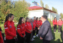 Görselde, yeşillikler içindeki bir açık alanda takım halinde duran ve kırmızı-beyaz spor kıyafetleri giymiş işitme engelli kadın güreşçi sporcular ile karşılıklı durarak onlarla konuşan takım yetkilisi veya antrenör bir erkek yer almaktadır. Arkada ise tek katlı bir bina ve ağaçlar bulunmaktadır.