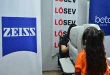 Görselde, turuncu tişört giyen bir çocuğun, optik mağazasında görevli personel eşliğinde özel bir cihazla göz muayenesi yaptırdığı; arka planda ZEISS, LÖSEV ve Beta Optik logolarının yer aldığı görülmektedir.