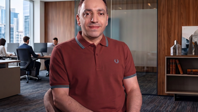 Mehmet Kızıltaş, ofis ortamında bir masada oturmuş şekilde kameraya doğru bakıyor. Üzerinde bordo bir polo tişört var. Arka planda cam bölmeli, modern dekorlu bir çalışma alanı ve bilgisayar başında çalışan iki kişi görülüyor. Ortam aydınlık, düzenli ve kurumsal bir ofis atmosferi taşıyor.