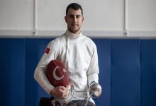 Türk bayrağı armalı beyaz eskrim kıyafeti giyen Hakan Akkaya, bir elinde ay-yıldızlı kılıfı, diğer elinde eskrim kılıcıyla spor salonunda poz veriyor.