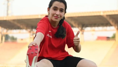 Görselin üst bölümünde, açık hava atletizm pistinde yeşil çim zemin üzerinde oturarak esneme hareketi yapan genç bir kadın sporcu yer almaktadır. Kırmızı tişört ve siyah şort giyen sporcu, bir eliyle ayağının ucundan tutarak esnerken diğer eliyle kameraya doğru baş parmağıyla onay (beğeni) işareti yapmakta ve gülümsemektedir. Arka planda stadyum tribünleri ve çatı yapısı görülmektedir.