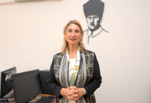 Kapalı bir mekânda, muhtemelen bir bilgisayar laboratuvarı ya da eğitim odasında duran Prof. Dr. Gülser Kılınç, kameraya doğru sakin ve kendinden emin bir ifadeyle bakıyor. Üzerinde siyah zeminli, altın ve beyaz zincir motifleriyle süslenmiş şık bir ceket bulunuyor. Boynunda açık renkli bir şal var; bu şal görünümüne zarif bir dokunuş katıyor. Arka planda, duvarda Mustafa Kemal Atatürk’ün siyah hat çizimiyle yapılmış portresi dikkat çekiyor. Çevrede masa üstü bilgisayarlar, klavyeler ve monitörler yer alıyor; bu da ortamın akademik veya kurumsal bir eğitim alanı olduğunu düşündürüyor. Prof. Dr. Gülser Kılınç fotoğrafta hem profesyonel hem de sıcak bir duruş sergiliyor; arka plandaki Atatürk portresi ise mekâna anlamlı bir kültürel vurgu katıyor.