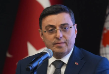 Koyu renk takım elbise giyen, gözlüklü Serkan Bayram mikrofonla resmi bir açıklama yapıyor. Arkasında Türk bayrağı bulunuyor. Ortam, ciddi ve kurumsal bir basın toplantısı ya da resmi etkinlik havası taşıyor.