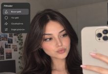 Bir genç kadın, elinde akıllı telefonuyla selfie çekerken burun şekli, yüz şekli, güzelleştirme ve doğal görünüm gibi sosyal medya filtrelerini gösteren bir arayüz eşliğinde yüz hatlarını inceliyor.