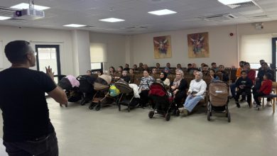 Görselde, bir salonda bir konuşmacının karşısında oturan kalabalık bir grup insan, çocuk arabaları ve çocuklarla birlikte dikkatle sunumu dinliyor; sağ tarafta ise bir kişi ayakta duruyor..