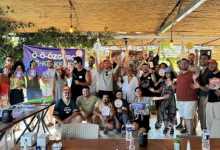 Geniş bir kapalı alanda, ahşap çatı ve sarkan yeşil bitkilerin altında büyük bir grup bir araya gelmiş. İnsanlar gülümseyerek kameraya bakıyor, çoğu ellerini havaya kaldırmış ve renkli kartlar, küçük pankartlar ya da etkinlik materyalleri tutuyor. Ortam canlı, neşeli ve bir kutlama havasında. Arka planda mor bir afiş üzerinde “Ö-Ö-ÖZGÜRCE K-K-KESFEET” yazısı görülüyor. Masalarda su şişeleri ve etkinlik malzemeleri var. Fotoğraf, kekemlik kampının enerjik, kapsayıcı ve dayanışma dolu atmosferini yansıtıyor.