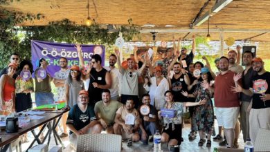 Geniş bir kapalı alanda, ahşap çatı ve sarkan yeşil bitkilerin altında büyük bir grup bir araya gelmiş. İnsanlar gülümseyerek kameraya bakıyor, çoğu ellerini havaya kaldırmış ve renkli kartlar, küçük pankartlar ya da etkinlik materyalleri tutuyor. Ortam canlı, neşeli ve bir kutlama havasında. Arka planda mor bir afiş üzerinde “Ö-Ö-ÖZGÜRCE K-K-KESFEET” yazısı görülüyor. Masalarda su şişeleri ve etkinlik malzemeleri var. Fotoğraf, kekemlik kampının enerjik, kapsayıcı ve dayanışma dolu atmosferini yansıtıyor.