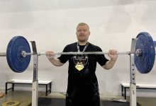 Görselde, spor salonunda halter barını kaldıran veya tutan, boynunda madalya takılı olan bir sporcu yer almaktadır.