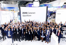 Kalabalık bir grup, aydınlık bir fuar alanında kurulan ZEISS Cataract Workflow standının önünde toplu halde poz veriyor. Katılımcıların çoğu iş kıyafeti ve yaka kartı taşıyor; ortam profesyonel bir kongre veya teknoloji etkinliği havasında. Arka planda modern tıbbi cihazlar ve parlak ışıklarla hazırlanmış sergi alanı görülüyor. Genel atmosfer, göz sağlığı ve katarakt teknolojilerine odaklanan kurumsal bir buluşmayı yansıtıyor.
