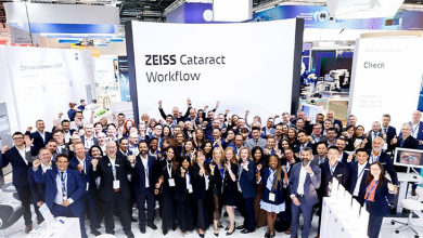 Kalabalık bir grup, aydınlık bir fuar alanında kurulan ZEISS Cataract Workflow standının önünde toplu halde poz veriyor. Katılımcıların çoğu iş kıyafeti ve yaka kartı taşıyor; ortam profesyonel bir kongre veya teknoloji etkinliği havasında. Arka planda modern tıbbi cihazlar ve parlak ışıklarla hazırlanmış sergi alanı görülüyor. Genel atmosfer, göz sağlığı ve katarakt teknolojilerine odaklanan kurumsal bir buluşmayı yansıtıyor.